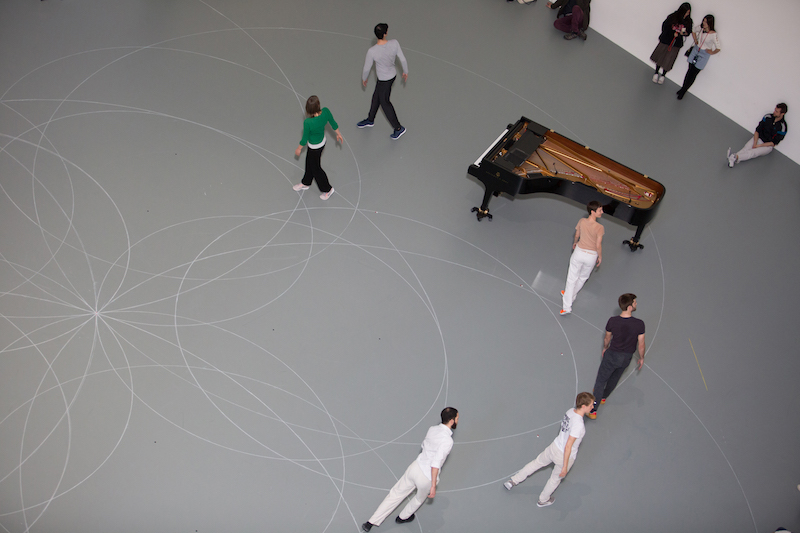 An aerial view of a group of dancers standing on one of the outlines of a circle. They all turn their heads over their shoulders. The piano is also set by them. 